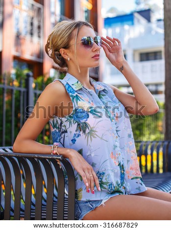 Cute blond woman in summer clothing posing over green bushes and building.