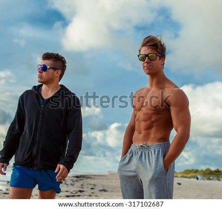 Two young muscular guys in sunglasses posing on a beach over blue sky with white clouds.
