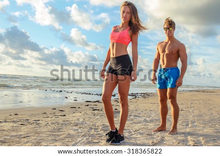 Muscular man and sporty woman standing on the beach over blue sky with clouds.