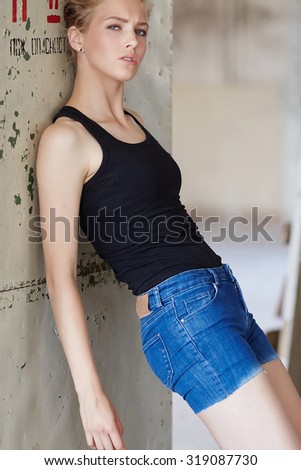 Awesome sexy blond girl in denim shorts and black shirt posing over grey wall.