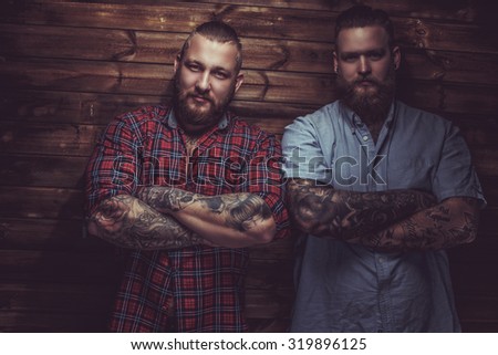 Two brutal mans with beards and tattooes on their hands posing over wooden wall.