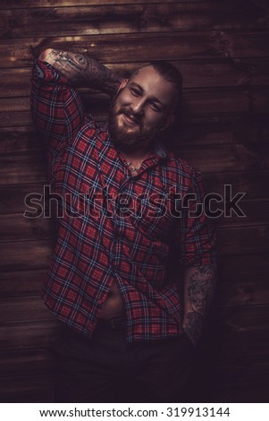Smiling man with tattooes and beard posing over wooden wall.