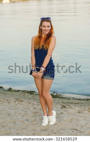 Redhead woman in jeans shorts and blue shirt posing on the beach near water.