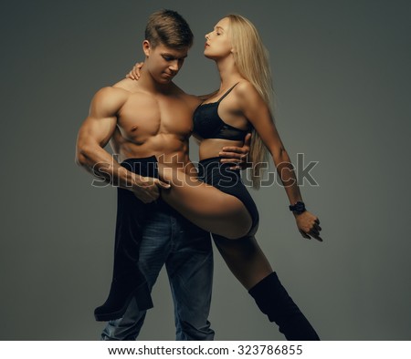 Shirtless muscular man holding long leg of his sexy blond woman. Isolated on grey background.