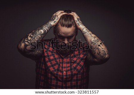 Brutal man with beard holding his head with tattooed arms and looking down. Isolated on grey background.