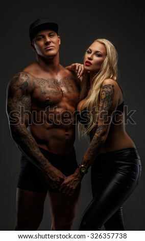 Strong shirtless tattooed man in a cap and blond sporty woman in black bra isolated on grey background.