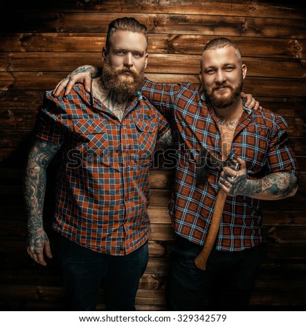 Two mans with beards and tattoo posing over wooden wall. One of them holding axe.
