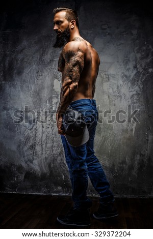 Full body portrait of muscular tattooed bearded man in blue jeans over grey wall.