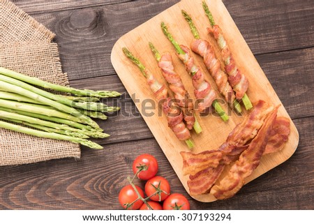 Asparagus wrapped in bacon with tomato on wooden background.