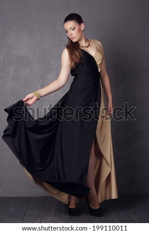 Young brunette lady in black ang golden dress posing on grey background. Fashion studio shot of beautiful woman with makeup and hairstyle wearing evening dress