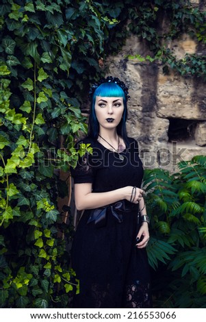 Beautiful goth girl with bright blue hair standing in the green ivy. On the eve of Halloween