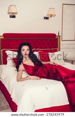 sexy glamour woman with black hair in elegant red dress in luxury bedroom