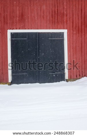 Barn doors, red wall, snow on the ground and light snow fall.