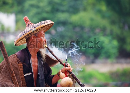Elderly Chinese fisherman in traditional clothes smoke cigaret.