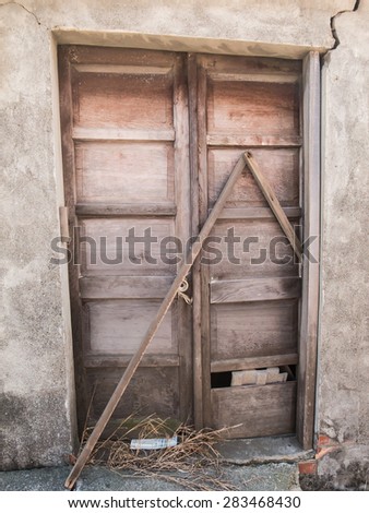 An old, broken wooden door. Two sticks blocking the door. A crack on the wall.