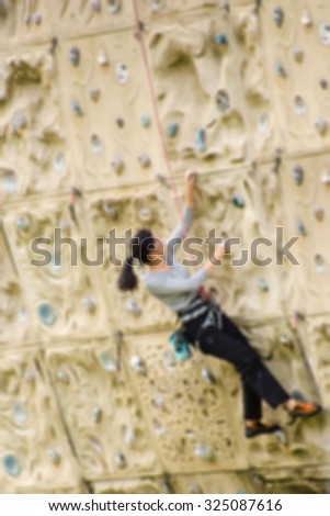 De focused/Blurred image of a young woman practicing rock climbing with tools hanging around her waist, a rope tied to her body. Young woman climbing up on practicing rock wall.