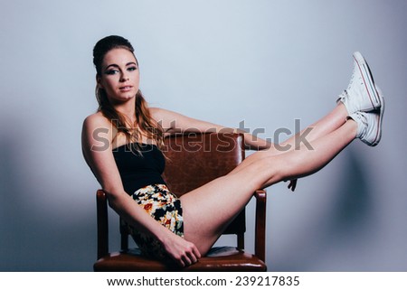 Studio Portrait of Young Attractive Woman sitting on brown leather chair sideways with legs sticking straight up
