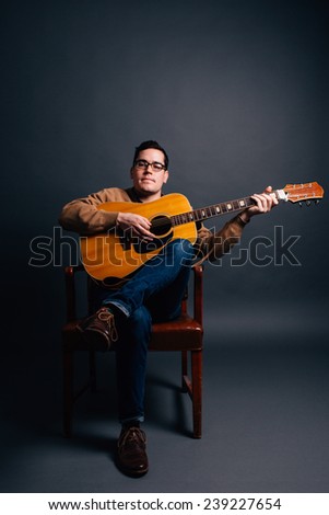 portrait of a young man in a sweater vest playing guitar sitting in chair