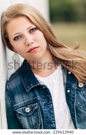 Attractive Young Blond leaning against white wall, looking at camera, no smile, jean jacket