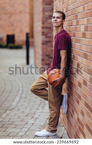 young male student holding basketball, slight smile, leaning against brick wall