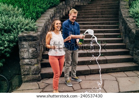 cute couple exploding a champagne bottle in front of stone steps laughing looking at camera