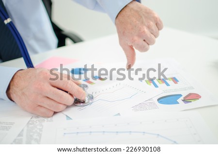Businessman before financial graphs with a stethoscope, thumb down