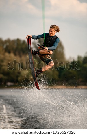 young man masterfully jumping over splashing wave on wakeboard. Sky and green trees in background Stock fotó © 