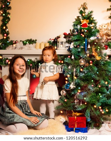 Girl with her little sister decorating Christmas tree sitting by the fireplace