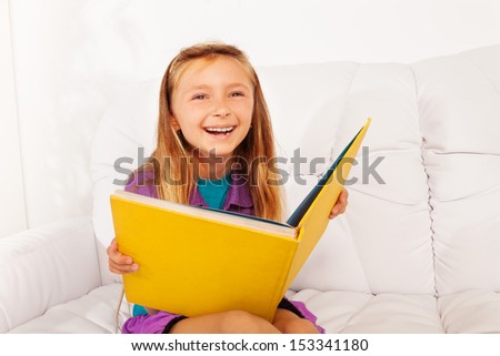 Laughing happy girl with book reading sitting at home on the sofa