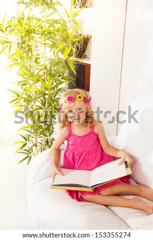 Nice Caucasian girl sitting on the couch with yellow book and reading wearing flowers wreath and smiling