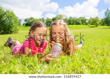 Two little girls lay in the grass looking at the red butterfly in the glass jar