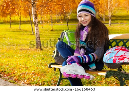 Close portrait of beautiful 11 years old girl putting on roller blades in warm clothes in the park sitting on the bench in autumn park with helmet laying on the bench
