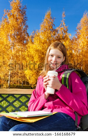 Beautiful teen 14 years old school girl sitting on the bench in autumn park holding coffee and textbooks
