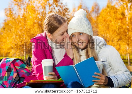 Two happy 14 years old girls whispering secrets and gossips in the autumn park sitting on the bench  holding textbook and coffee