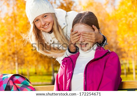 Two happy 14 years old girls in the autumn park playing guess who with one girl covering face with palms and laughing