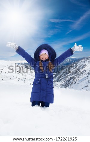 Happy 8 years old Caucasian girl in blue sitting with lifted from excitement hands on snow outside in winter