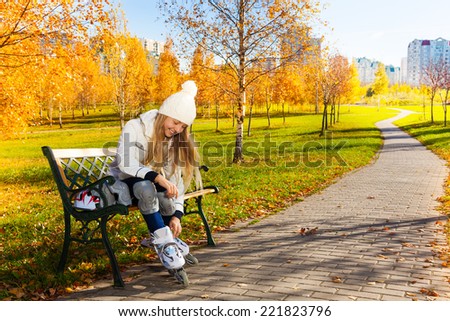 Blond teen girl with long hair in autumn park sitting on the bench and putting on roller blades