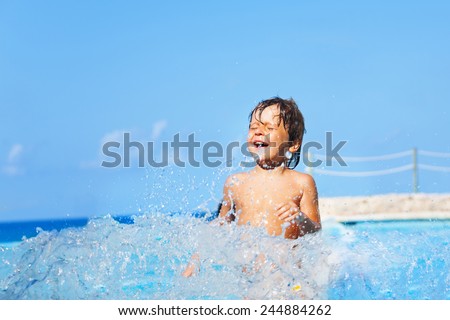 Laughing boy plays and splashes water around him in swimming pool with seashore background