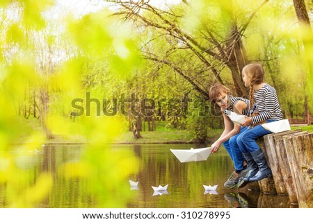 Boy and girl together near the pond play with paper boats on the water in beautiful forest landscape