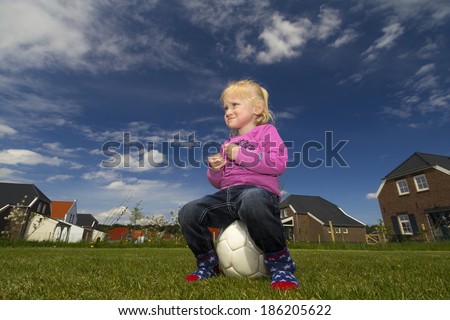 A little blonde girl with blue eyes, pink sweater and wearing no shoes, sits on a soccer ball.