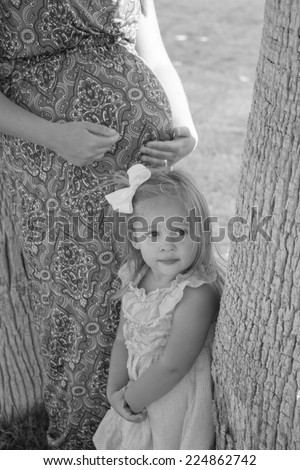 Woman with pregnant belly and toddler girl in black and white resting against trees in the shade