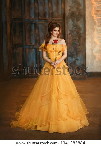 Girl beauty fantasy princess in yellow long historical, medieval silk dress holding flower red rose in her hands. Background of old gothic castle room. Fairy tale bewitched queen. Happy woman smiles.