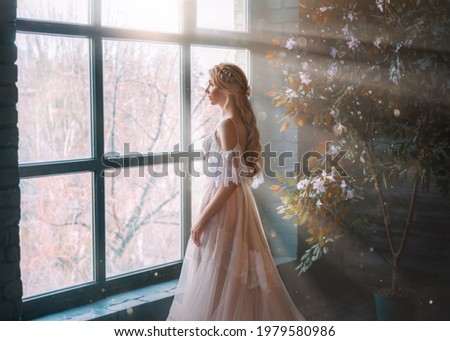 Romantic lady, blonde woman with long hair in white vintage dress stands in dark room, looks out window. Girl bride princess in wedding dress. Elegant hairstyle. Bright rays of sun concept of waiting.