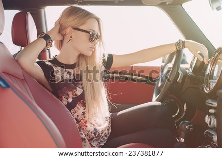 Blondie young girl at the wheel of sport car with red interior with black sunglasses and leather armlets with metal inserts, seating sideward with hand near her head and looking at the road