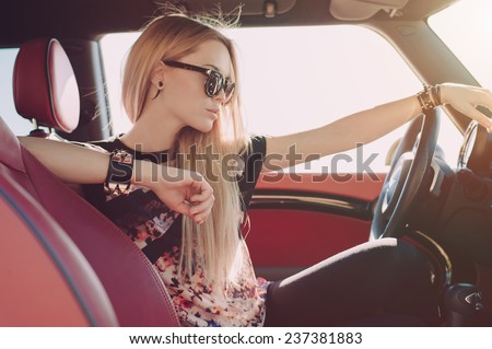 Blondie young girl at the wheel of sport car with red interior with black sunglasses and black leather armlets with metal inserts seating sideward and looking at the road
