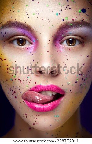 Close up facial beauty portrait of young funny girl with a lot of blur of pain on her face, pink lipstick, purple eyeshadows looking at you and sticking out her tongue on black background