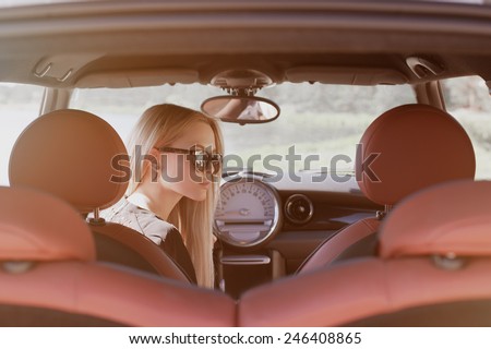 Nice portrait of blonde young woman at the wheel of sport car with red interior, with sunglasses, natural makeup looking to the right