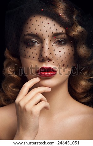 Facial portrait of lady with brown eyes, deep red lipstick, two nevus on her left cheek, curly hair and veil with black dots and rubies. Touching her lips. Black background.