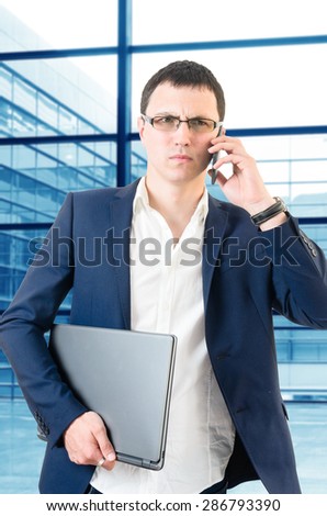 Young business man talking on the phone looking angry over big office's windows background