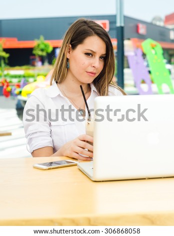 Smiling Young Woman Working on Laptop and speaking on the Smart Phone at Shopping Mall while Drinking Juice